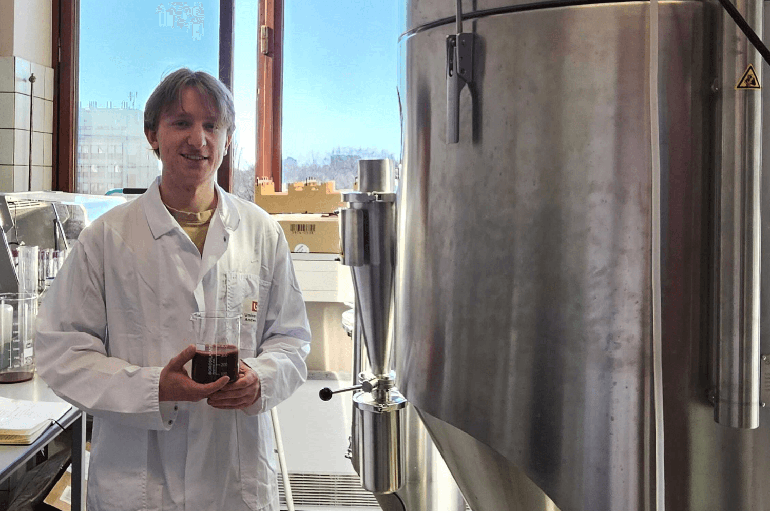 A person in a white lab coat stands in a laboratory next to a large stainless steel industrial tank, holding a container with dark red liquid, with daylight coming through a window behind them.