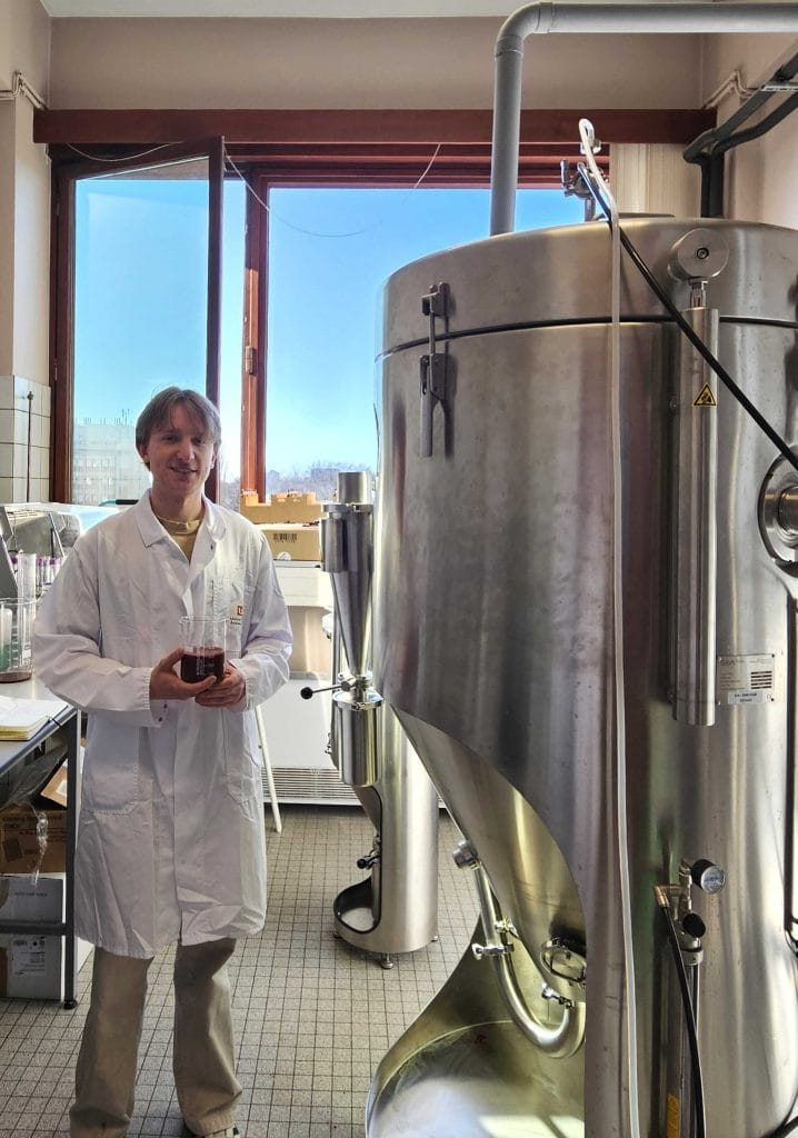 A person in a white lab coat stands in a laboratory next to a large stainless steel industrial tank, holding a container with dark red liquid, with daylight coming through a window behind them.