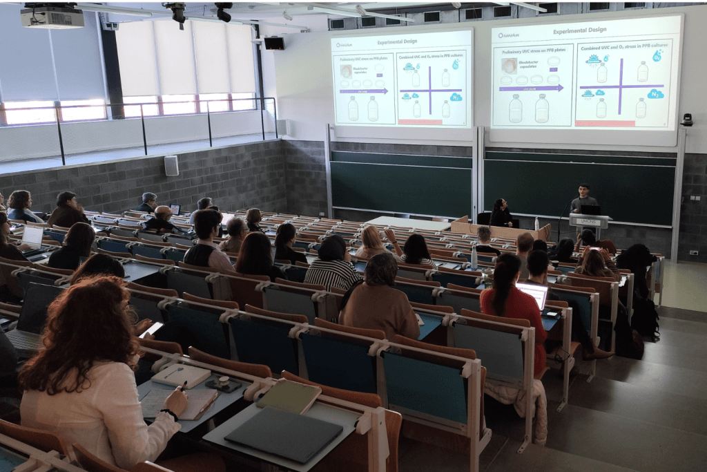Audience attending a lecture in a university auditorium with a presenter and slides displayed on large screens.
