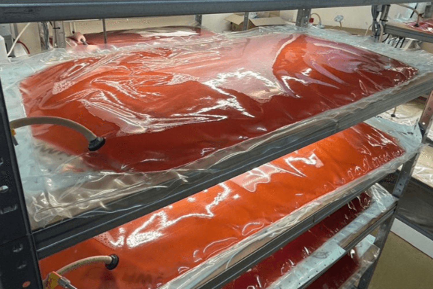 Multi-level metal shelving system in a laboratory holding several transparent plastic bioreactor bags filled with red-orange liquid culture. Tubing and monitoring equipment are connected to the bags, which are illuminated by overhead lights in a controlled indoor research environment.