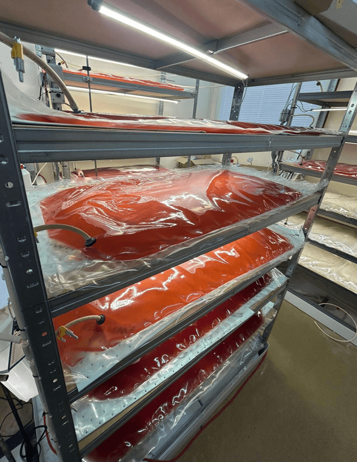 Multi-level metal shelving system in a laboratory holding several transparent plastic bioreactor bags filled with red-orange liquid culture. Tubing and monitoring equipment are connected to the bags, which are illuminated by overhead lights in a controlled indoor research environment.