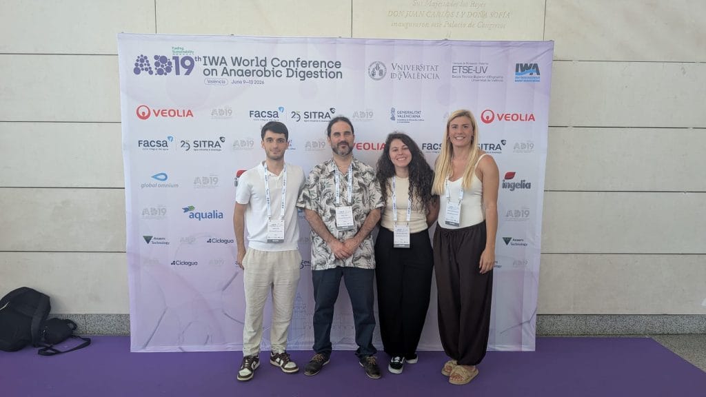 Four researchers wearing conference badges pose in front of the official backdrop at the 19th IWA World Conference on Anaerobic Digestion in Valencia. The backdrop features the conference title along with the logos of the University of Valencia, the International Water Association (IWA), and event sponsors.